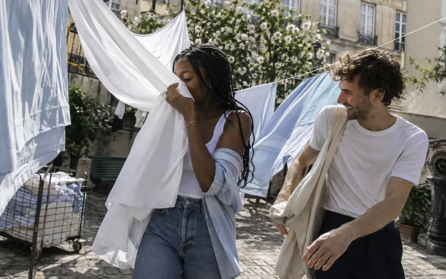 Mulher negra sente o cheiro de um lençol ao lado de um homem num pátio com roupas ao vento